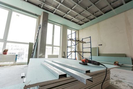 Construction site interior showing metal studs and drywall panels with a power tool on a stack of boards.