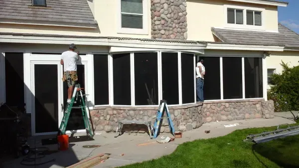 Two men are working on a screened in porch in front of a house.
