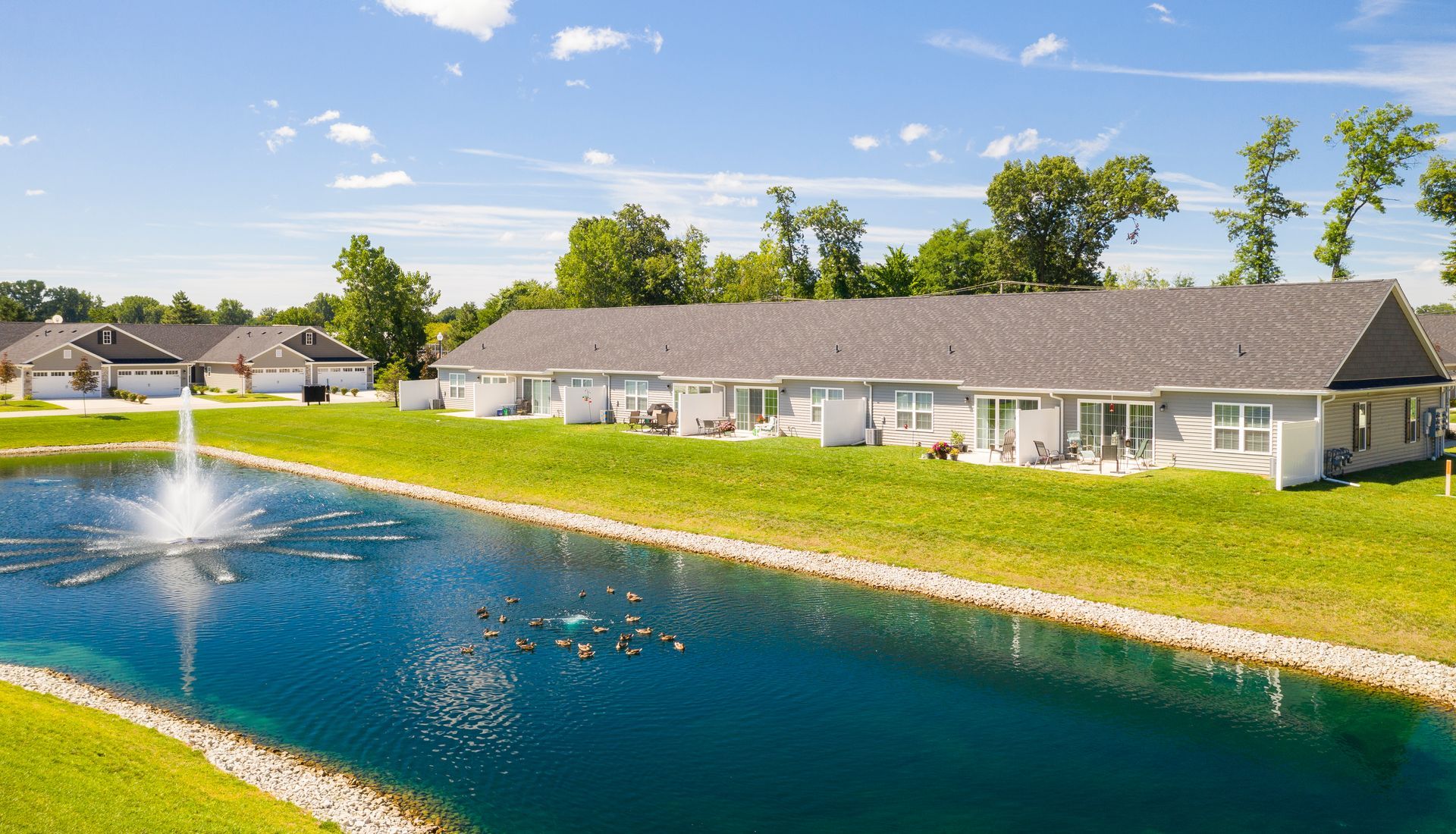 Apartment buildings beside a blue pond with a fountain and green lawns under a sunny sky