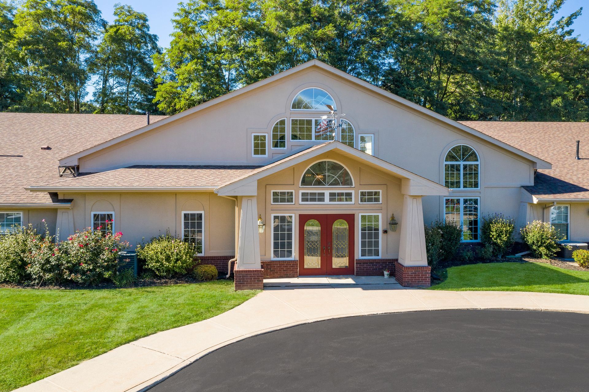 Front view of a beige house with a red double door, arched windows, and a curved driveway.