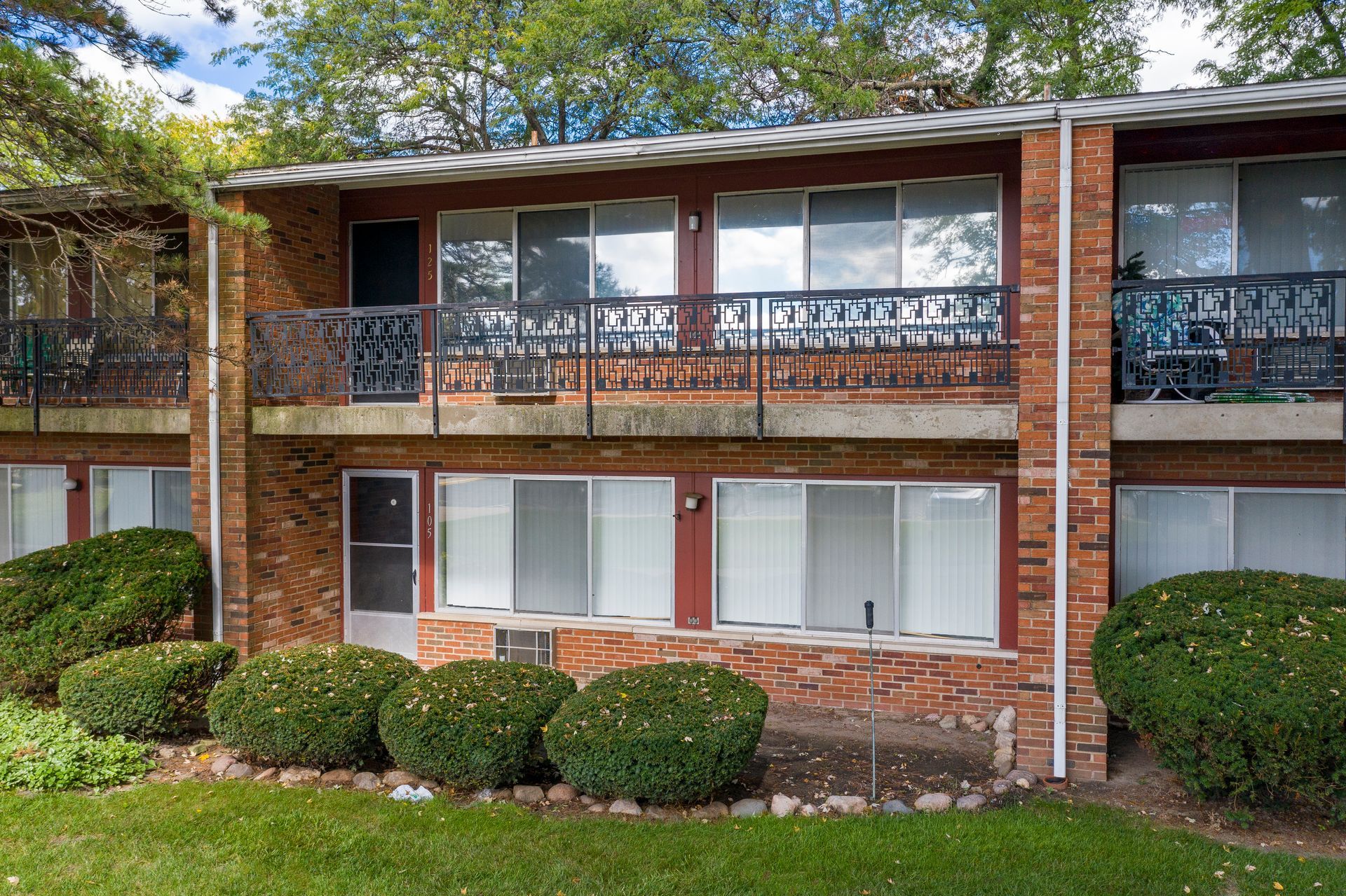 Brick apartment building with black balcony railings, white trim, and trimmed bushes in front