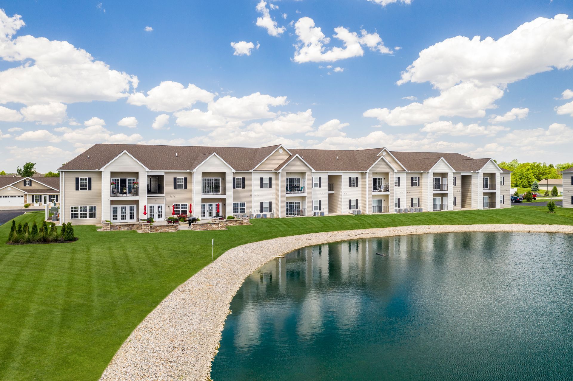 Apartment complex beside a landscaped pond under a blue sky with scattered clouds