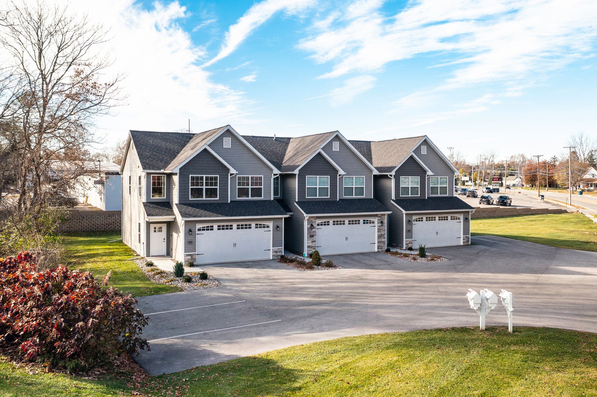 Three gray townhouses with garages in a quiet suburban neighborhood under a bright blue sky