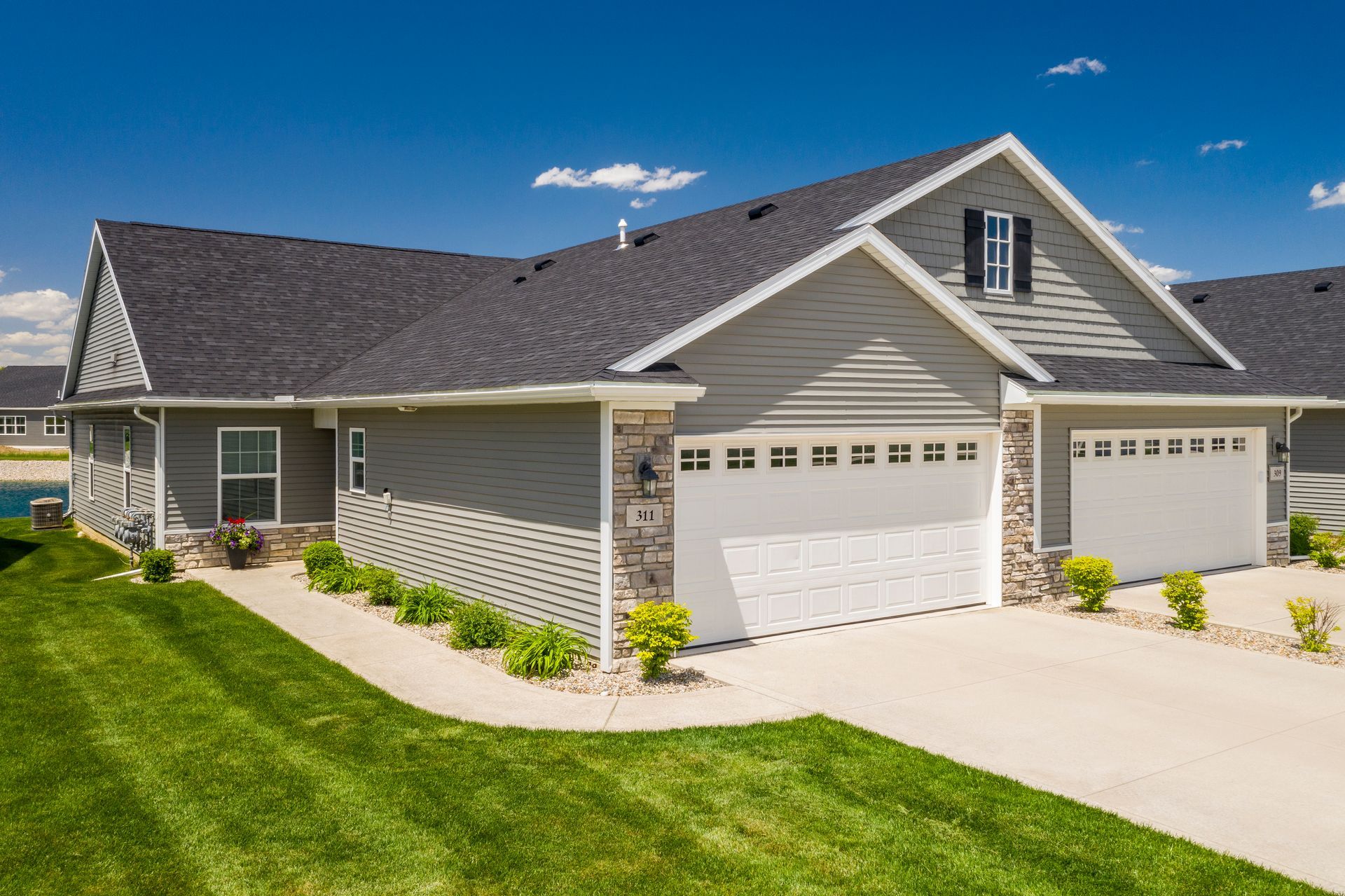 Suburban single-story house with gray siding, white garage doors, and a green lawn under a blue sky