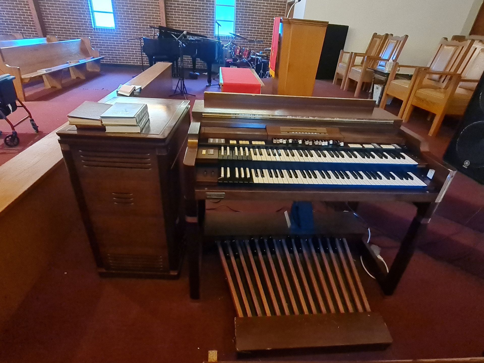 A Hammond organ in a church setting with wooden pews and other musical instruments.