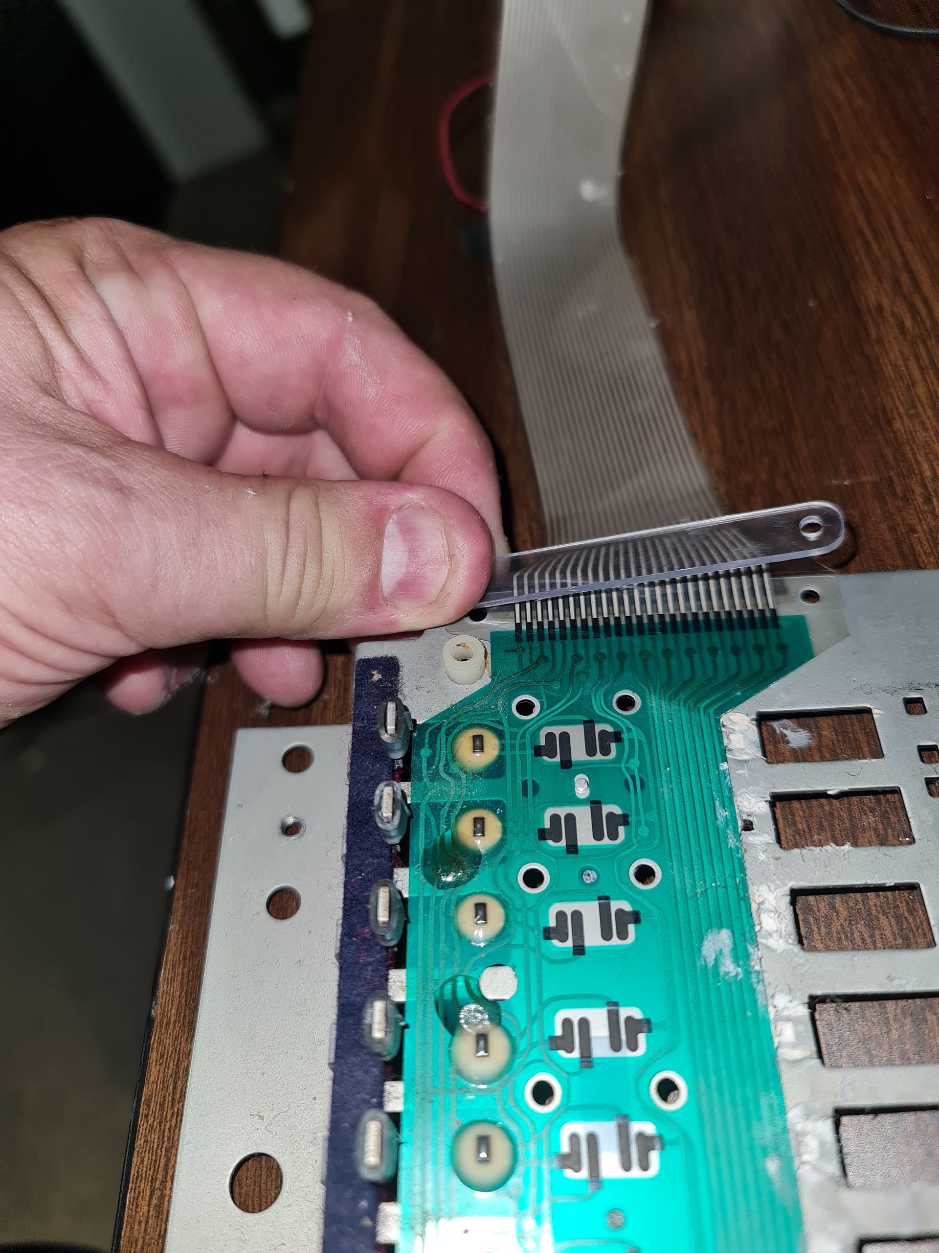 Hand holding a ribbon cable connected to a green circuit board with round components; table in the background.
