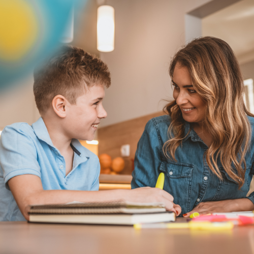 Woman helping a boy with homework, smiling, in a kitchen setting. They are looking at each other.