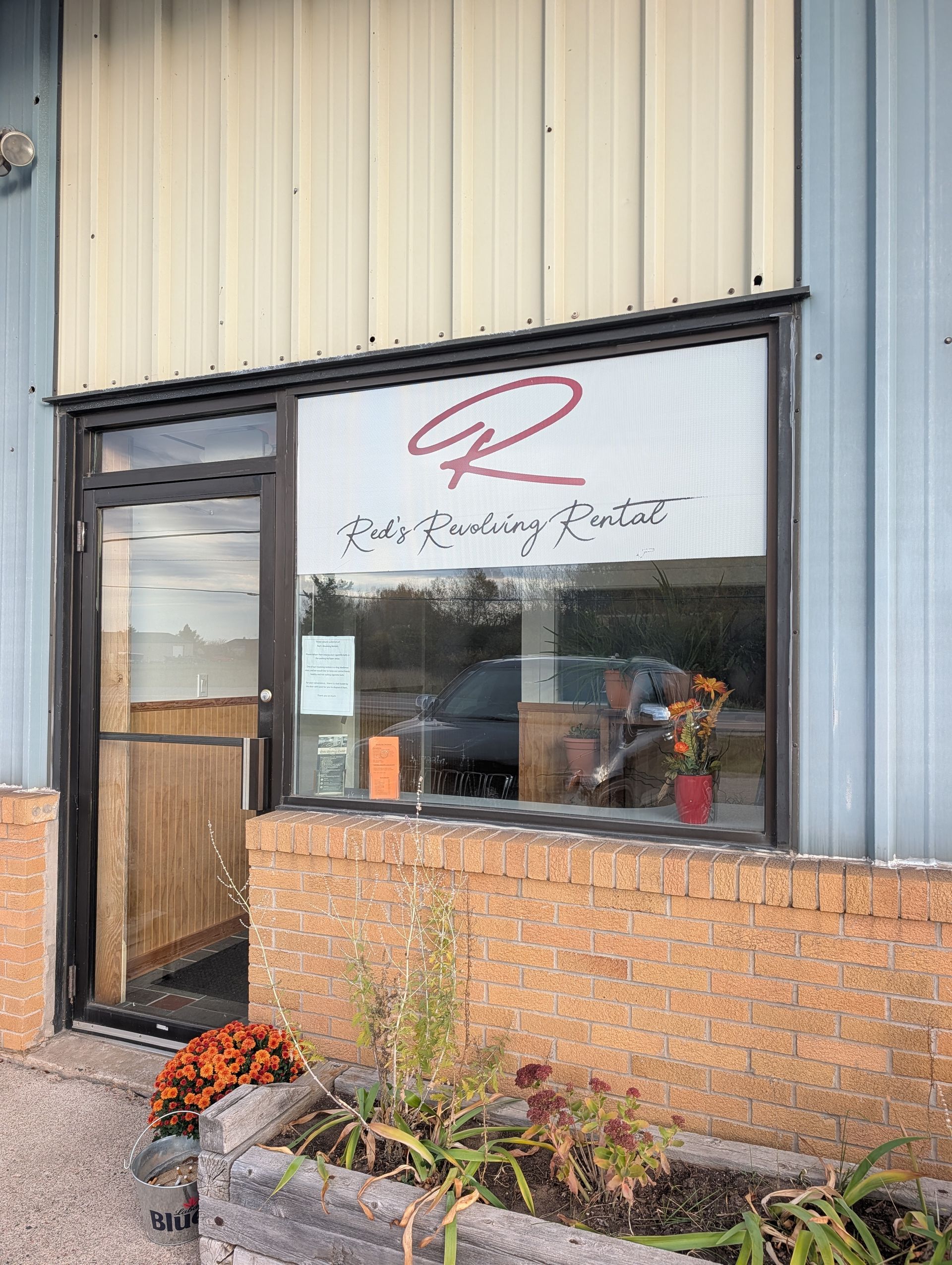 Storefront window with "Rudy's Roofing" sign, brick wall, and potted plants outside.