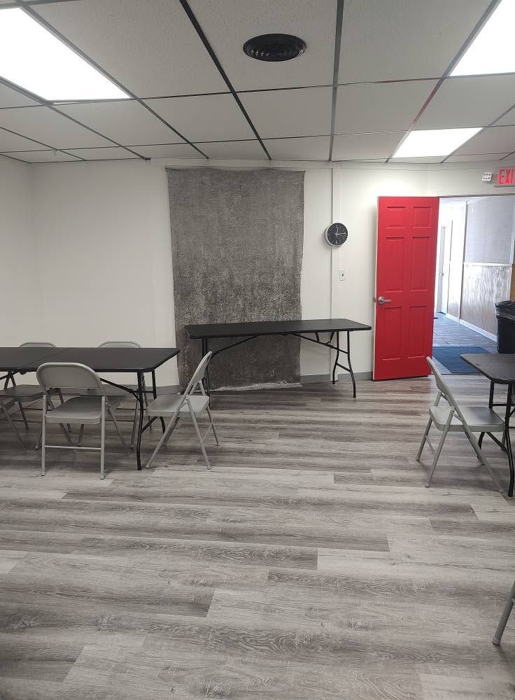 Empty room with folding tables and chairs, red door, and fluorescent ceiling lights.