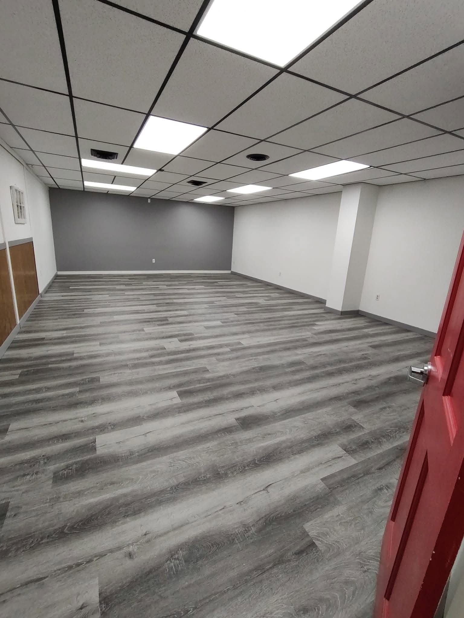 Empty office room with gray wood floor, drop ceiling, and white walls, viewed from an open doorway