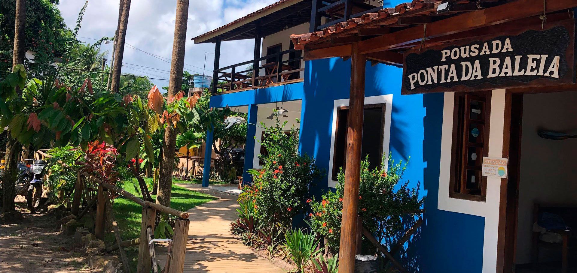 Um edifício azul vibrante com uma placa de madeira onde se lê "Pousada Ponta da Baleia", rodeado por árvores e um caminho de jardim.