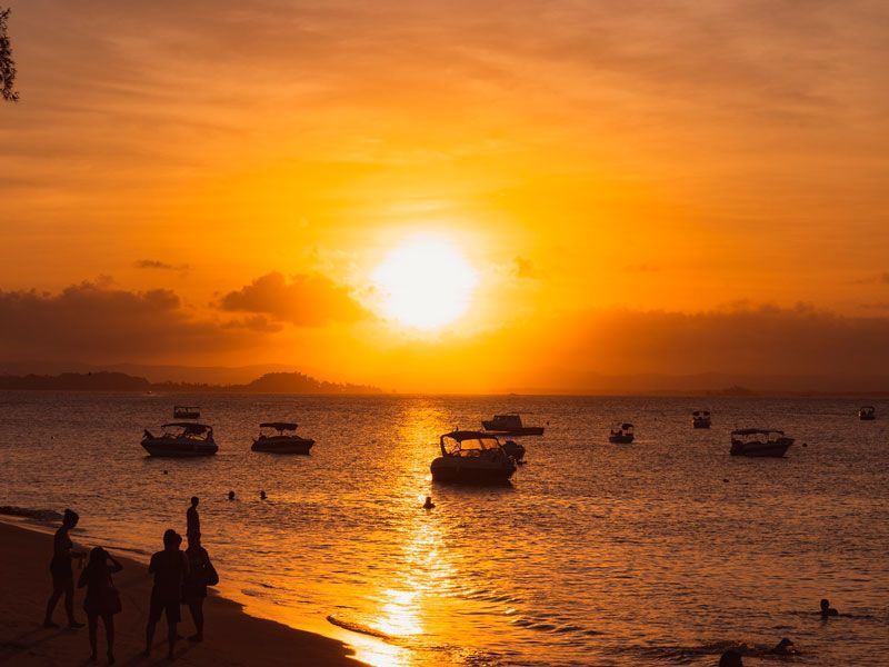Silhuetas em uma praia observam barcos flutuando na água durante um vibrante pôr do sol alaranjado.