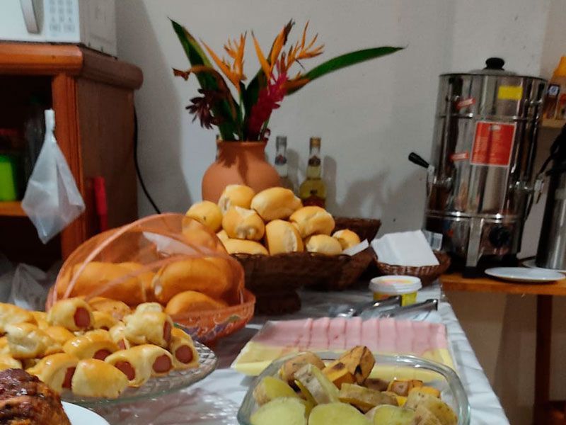 Um buffet de café da manhã com pãezinhos, salsichas enroladas em massa folhada, presunto fatiado, queijo e uma grande cafeteira.