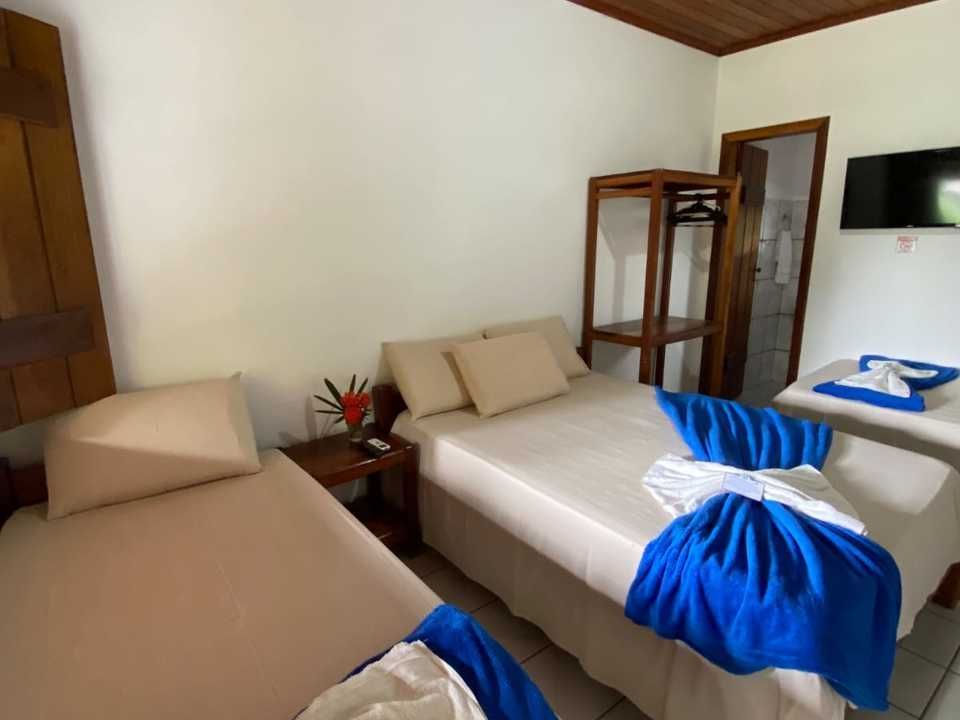 Um quarto de hotel com duas camas, roupa de cama bege, detalhes em azul, um cabideiro de madeira e uma TV fixada na parede.
