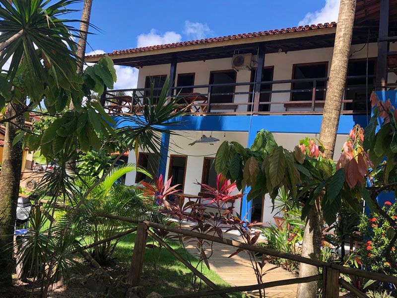 Um prédio de dois andares com fachada azul, varanda e telhado de telha, cercado por vegetação tropical exuberante e palmeiras.