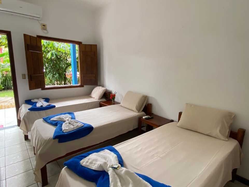Um quarto de hotel com três camas de solteiro, cada uma com roupa de cama bege e uma toalha azul dobrada por cima, perto de uma janela aberta.