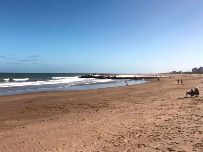 Uma praia ensolarada com um quebra-mar que se estende mar adentro, areia fina, ondas suaves e algumas pessoas relaxando ao longe.