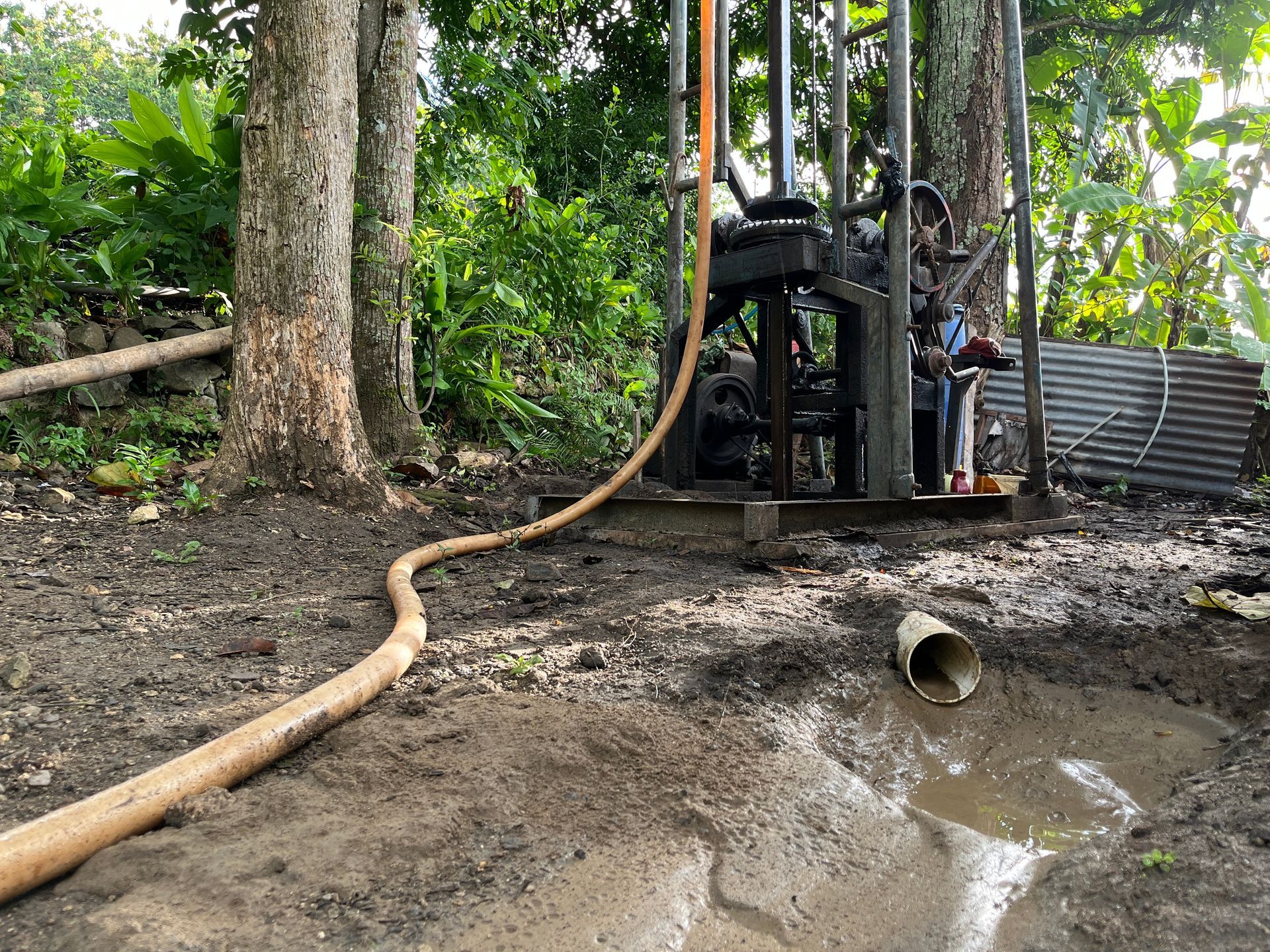 A drill rig operates in a muddy, wooded area, with a long hose extending toward a drainage pipe in a shallow pool.
