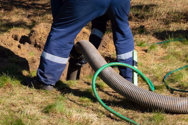 Person in blue work suit near a hole with a hose, possibly working on septic system outdoors.