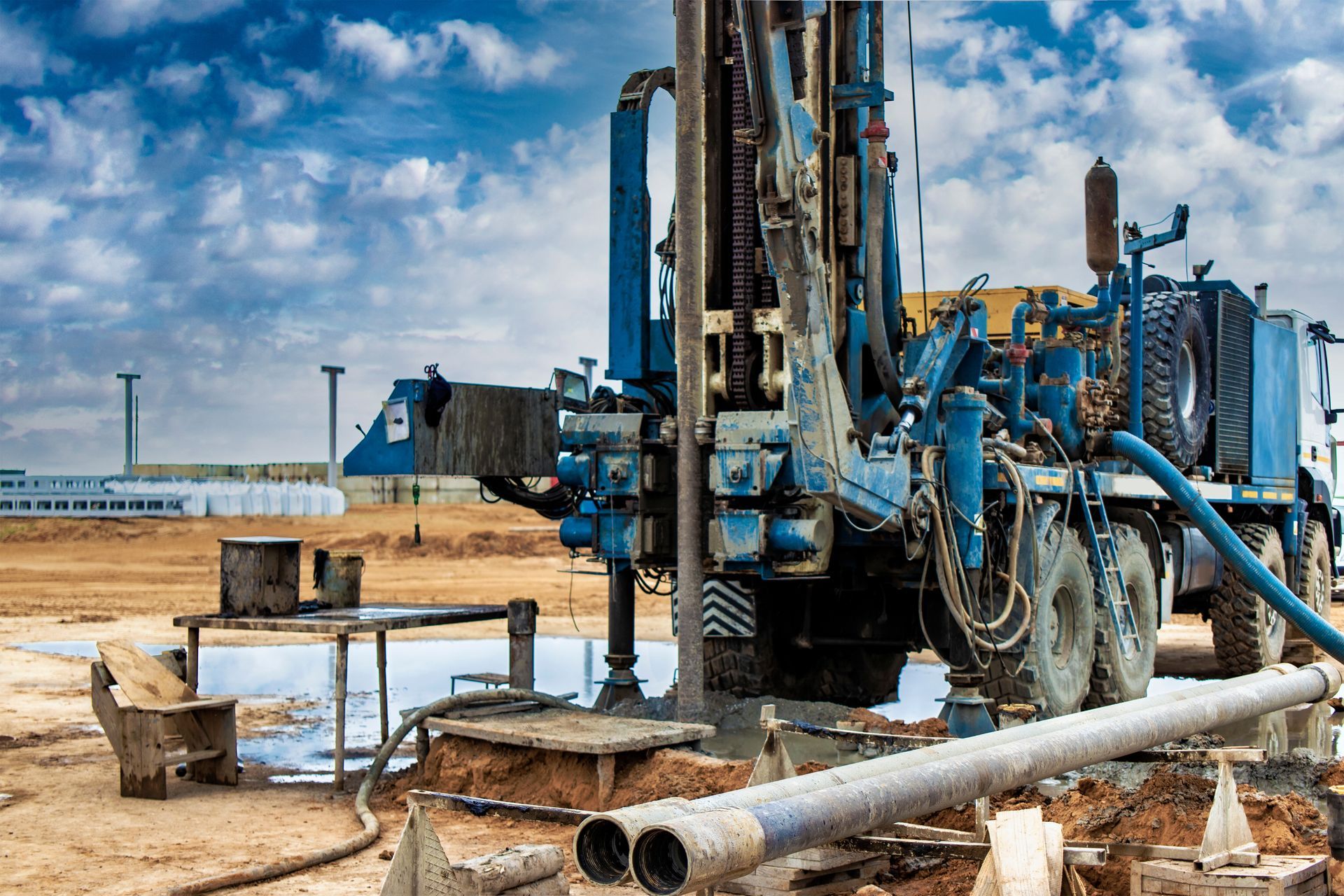 Blue drilling rig on a construction site, drilling into the ground, water present, under a cloudy sky.
