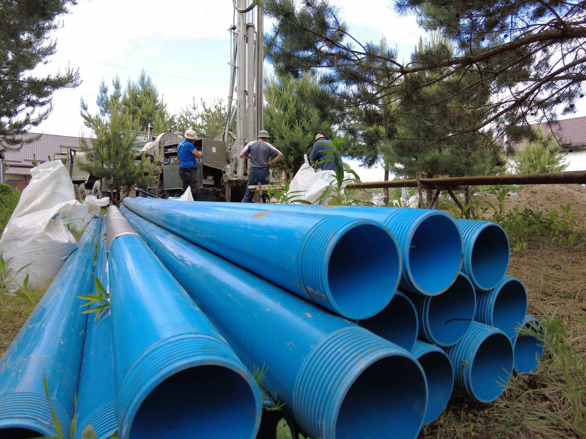 Blue PVC pipes stacked outdoors near a well drilling rig; workers in the background.