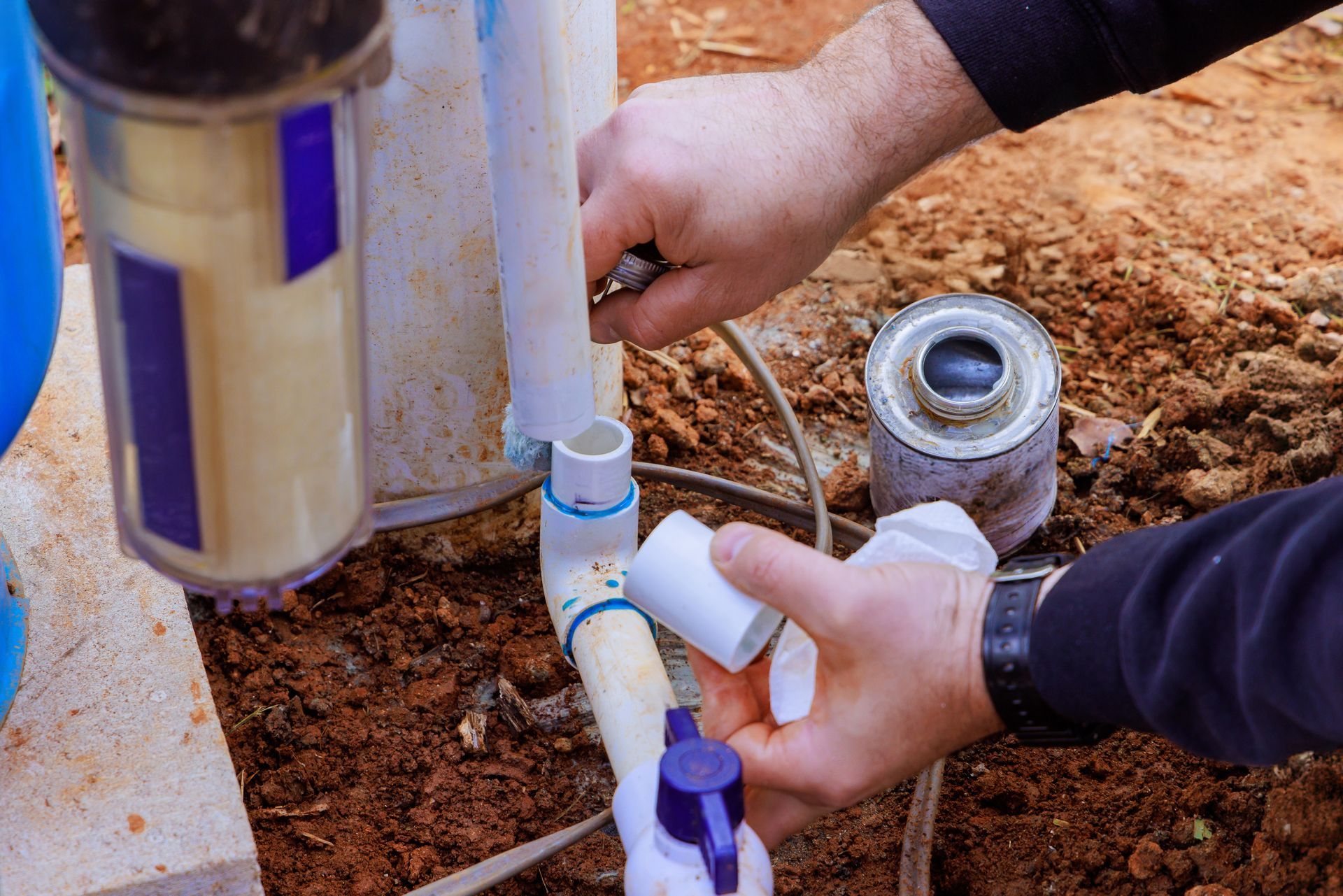 Hands connecting PVC pipe fittings, with glue can in a dirt setting.