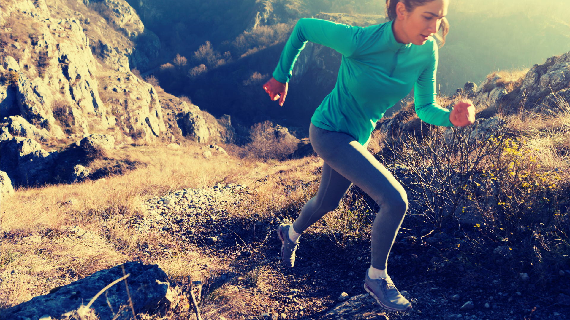 Woman running uphill on a rocky trail, wearing teal shirt and gray leggings.