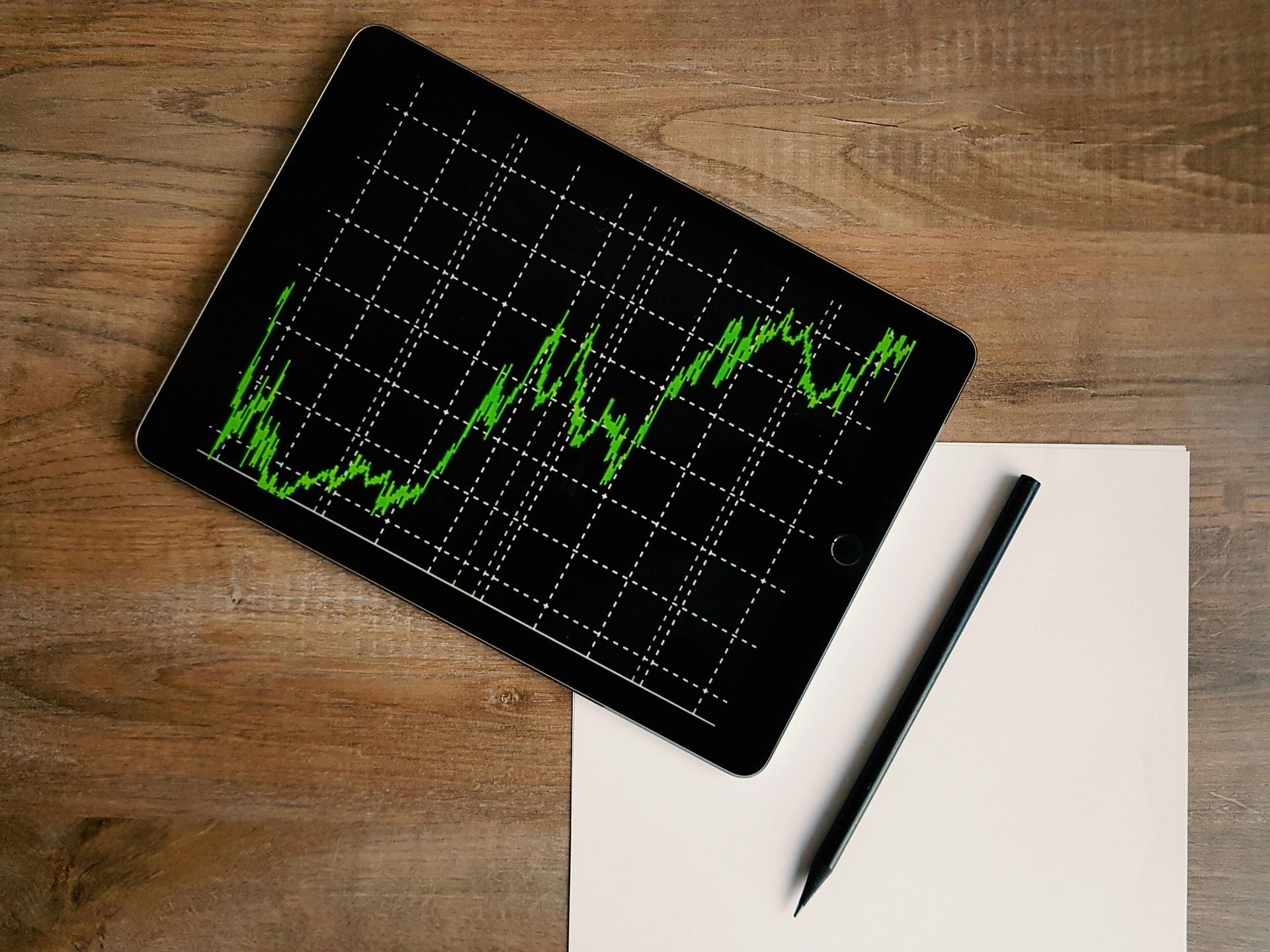 A tablet with a stock graph on it is sitting on a wooden table next to a pen and a piece of paper.