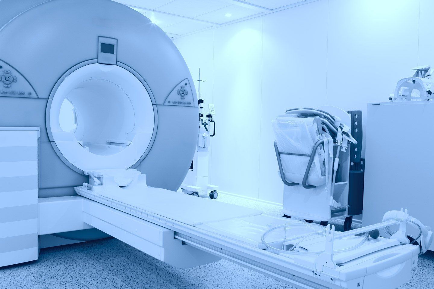 Una risonanza magnetica in una stanza medica con un letto per pazienti esteso di fronte ad essa, mostrata in una fredda tonalità monocromatica blu.