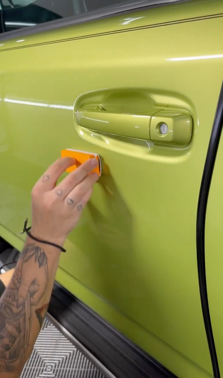 A person is polishing the door handle of a green car.