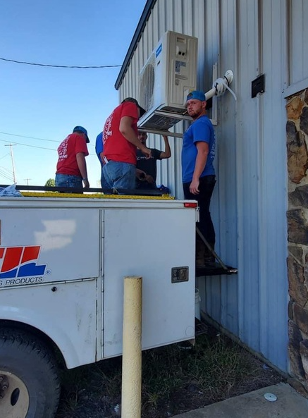 HVAC technicians installing an outdoor AC unit on a building. White truck parked nearby.