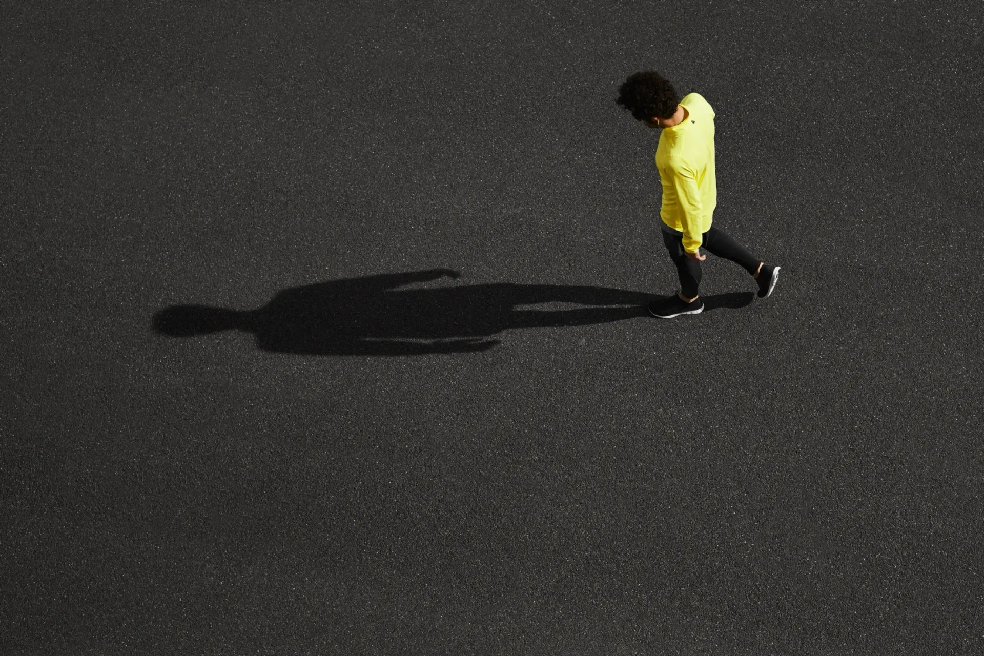 Person in a yellow coat walking on dark pavement, casting a long shadow.