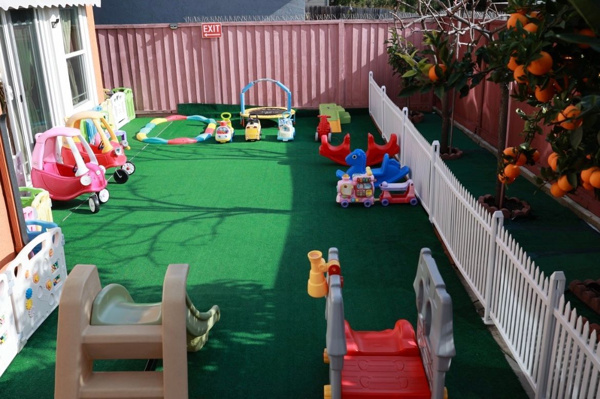 Playground with colorful toys on green turf, orange tree, picket fence, and wooden fence.