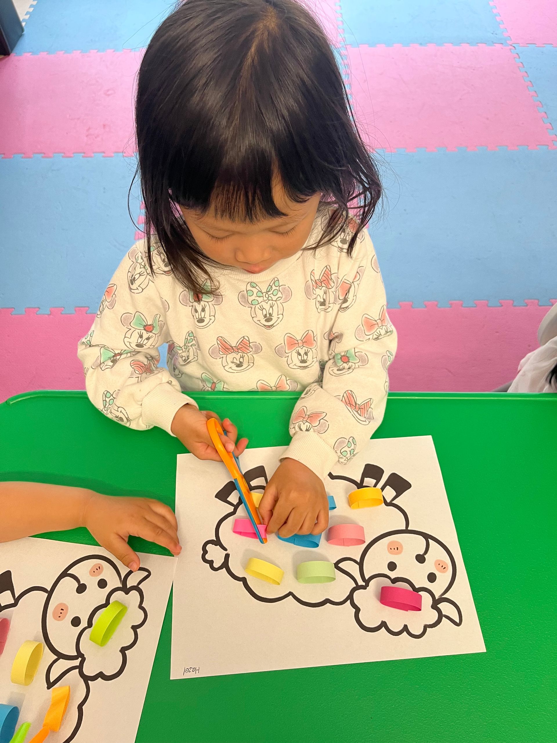A child coloring a drawing of a character using colorful clay on a green table.