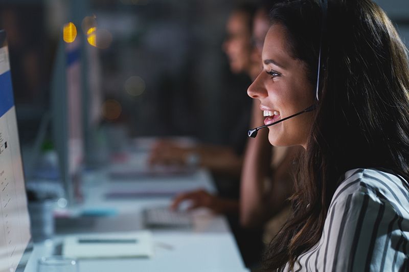 Customer support agent wearing a headset smiles at a computer in a dim office.