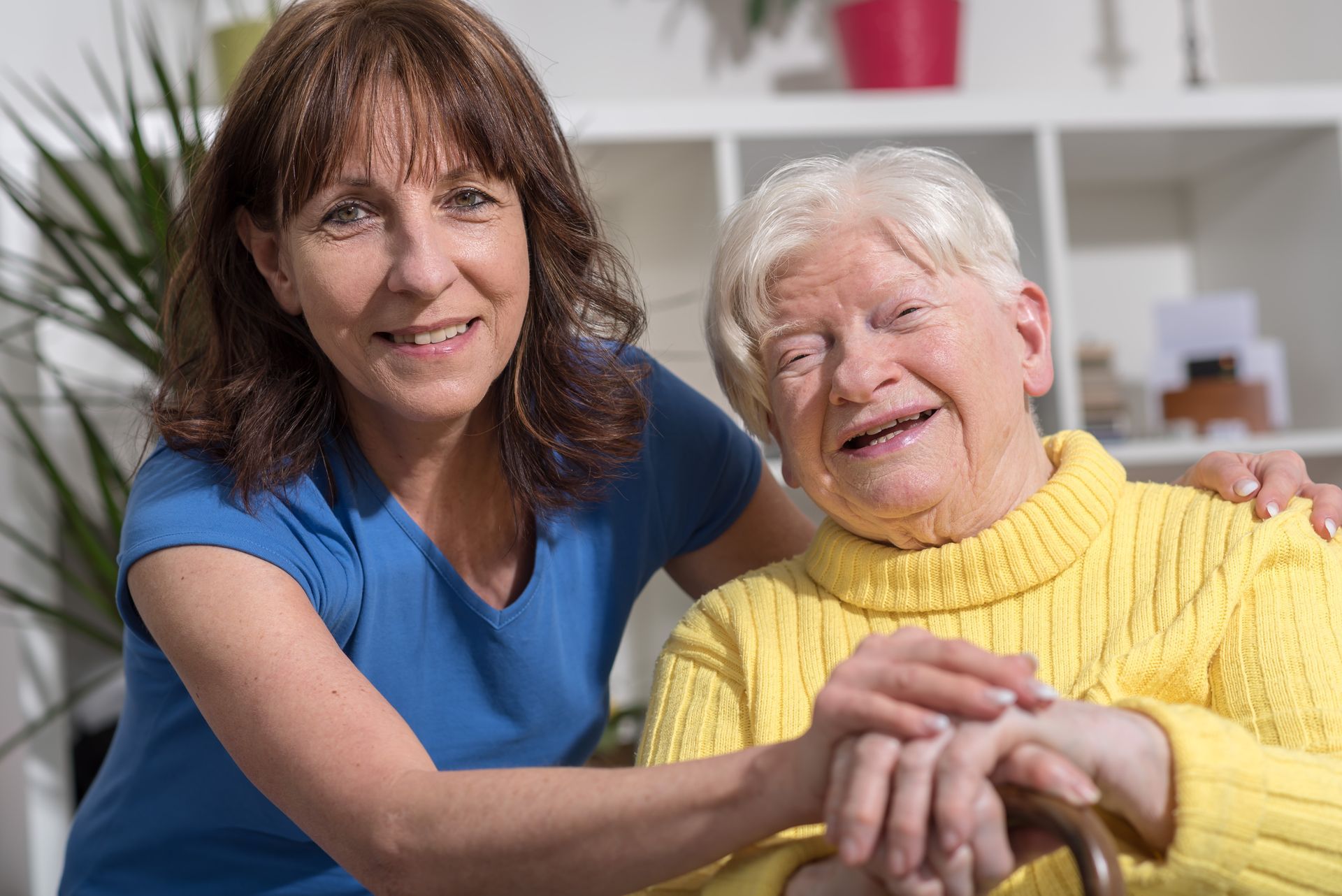 Caregiver smiling with an elderly woman in a yellow sweater, seated together indoors