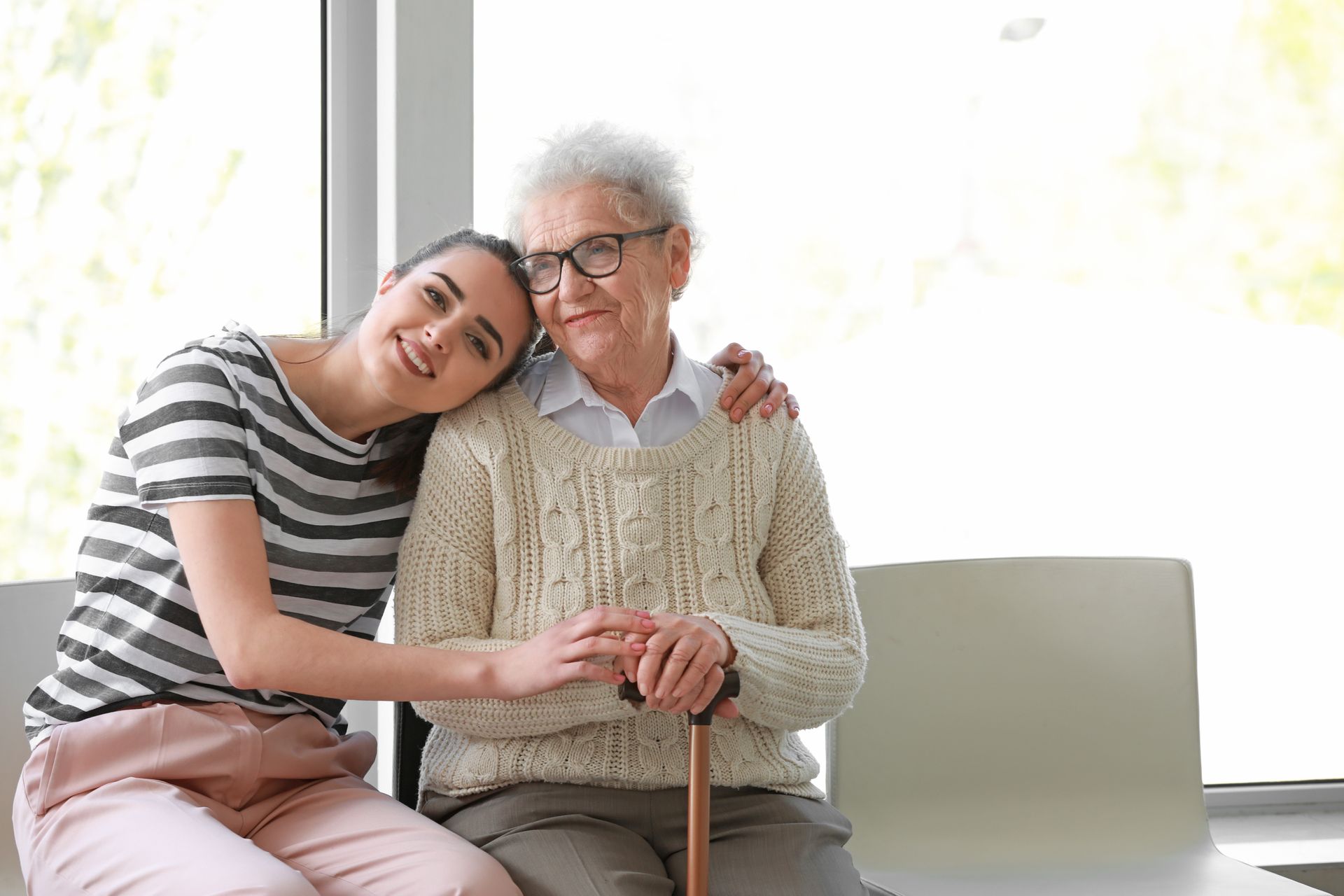 Young adult leaning on an elderly person on a sofa, both smiling in a bright room