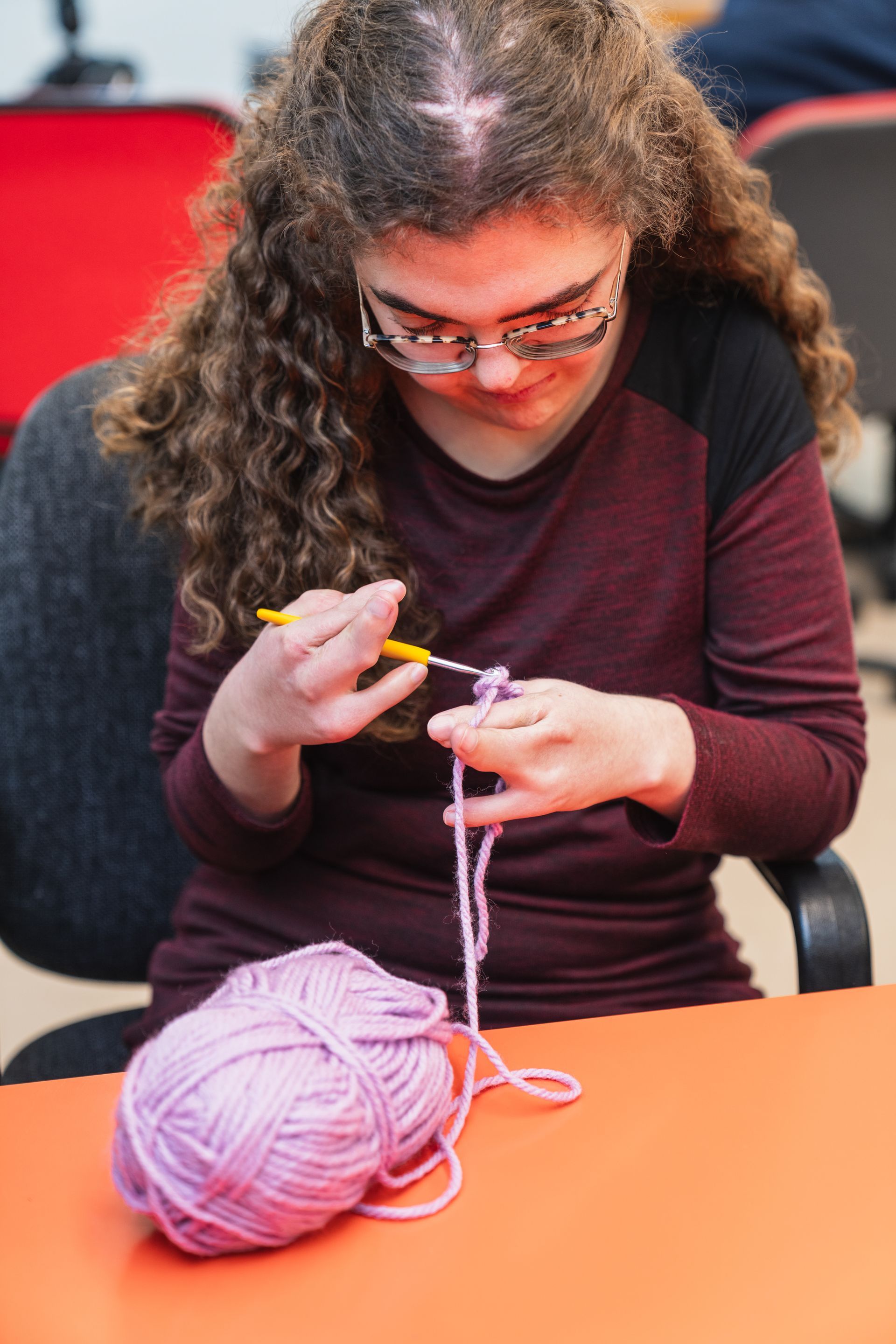 Person knitting with pink yarn at an orange table, concentrating on a small project.