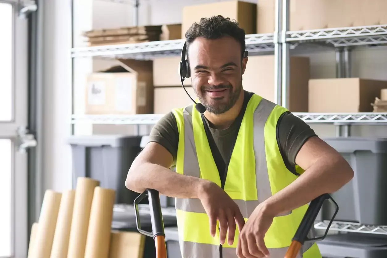 Smiling warehouse worker in a yellow safety vest leaning on a hand truck in a storage room