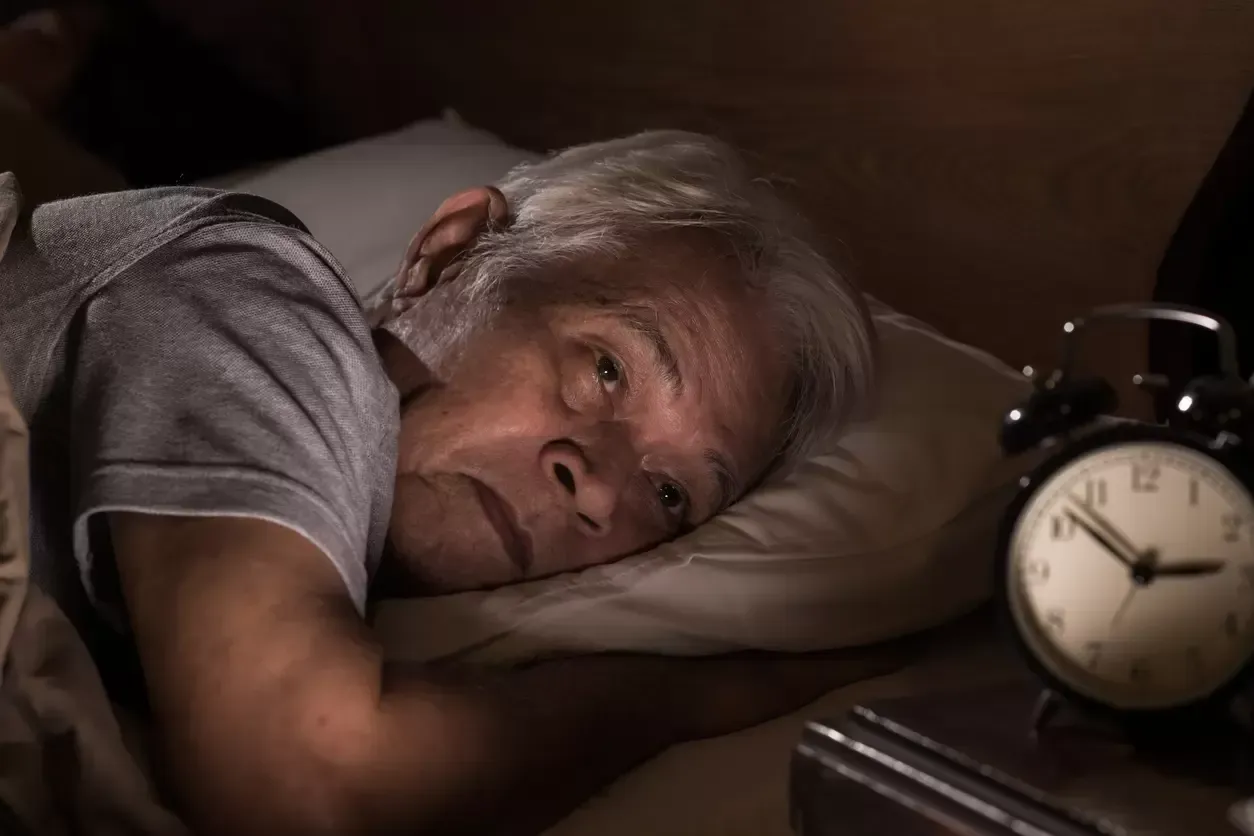 Elderly person lying in bed, looking at a glowing bedside alarm clock in a dim room