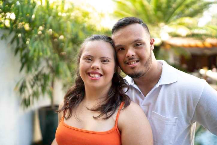 Smiling couple standing outdoors in front of greenery, with the woman in an orange top and the man in a white shirt