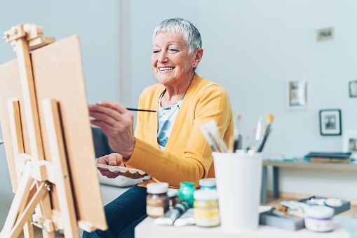Artist painting at an easel in a bright studio, smiling while working with brushes and paint jars.