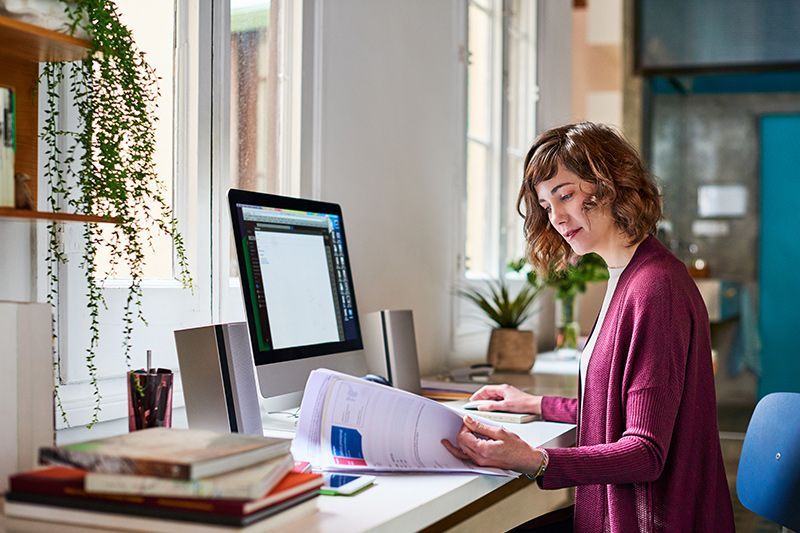 Person working at a desk with papers and computer in a bright office, holding documents and looking down
