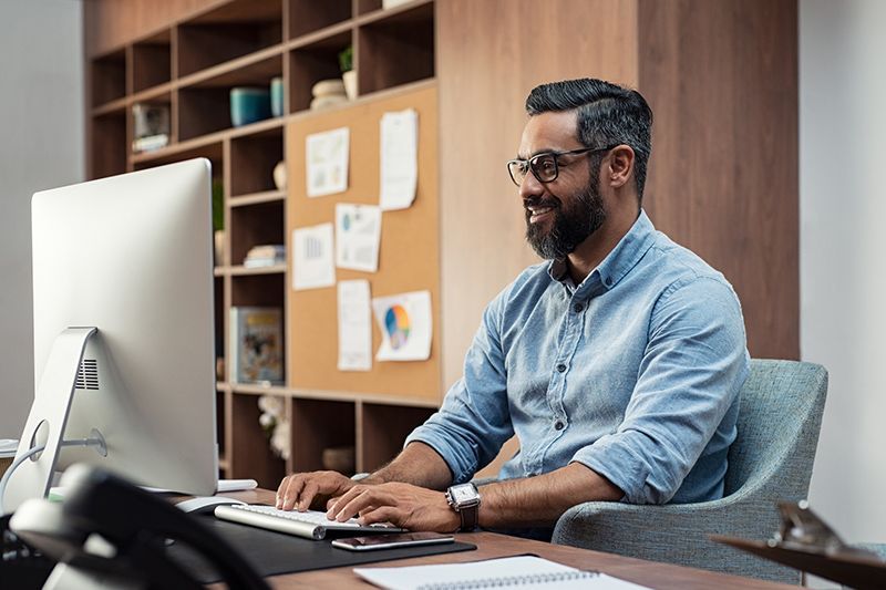 Man working at a desk in an office, typing on a computer with shelves and notes behind him.