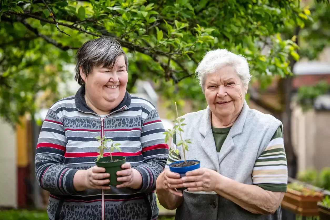 Two smiling people holding potted plants in a green garden outdoors