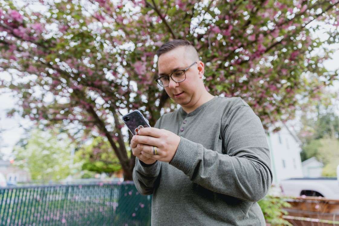 Man in gray sweater looking at a phone outdoors beneath pink flowering trees