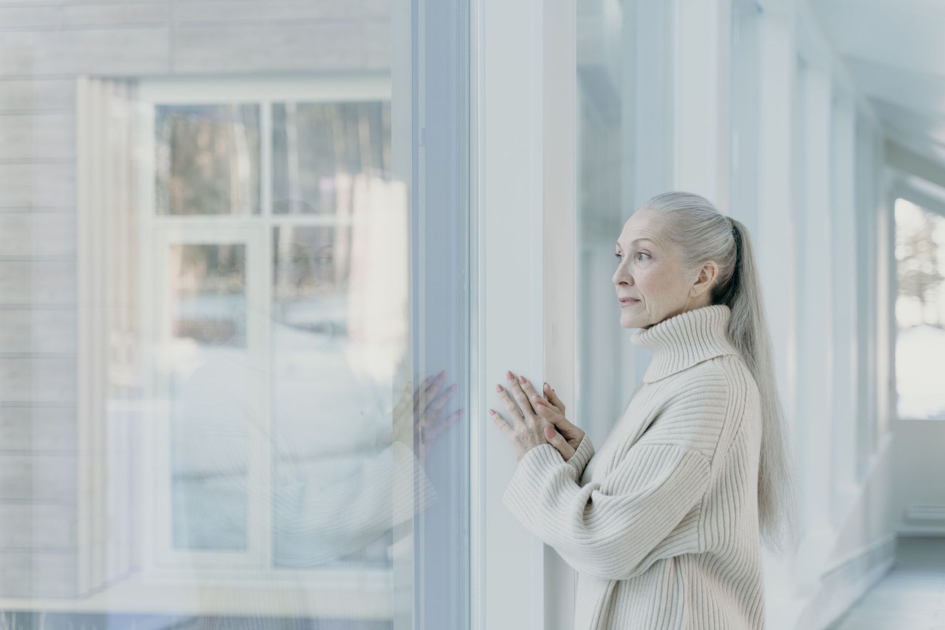 An older woman standing at a bright window in the late afternoon, looking out