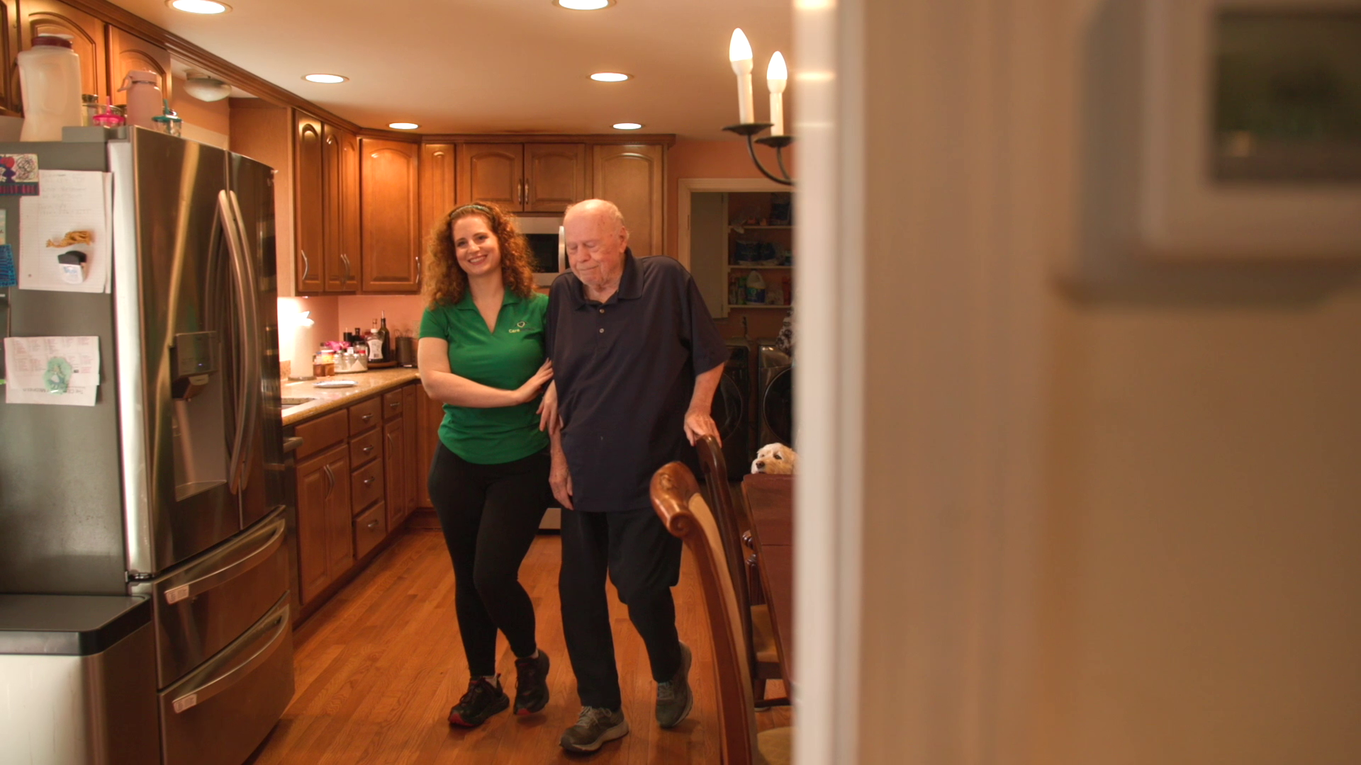 Two people standing together in a warm-lit kitchen, one in a green top and one in a dark shirt.