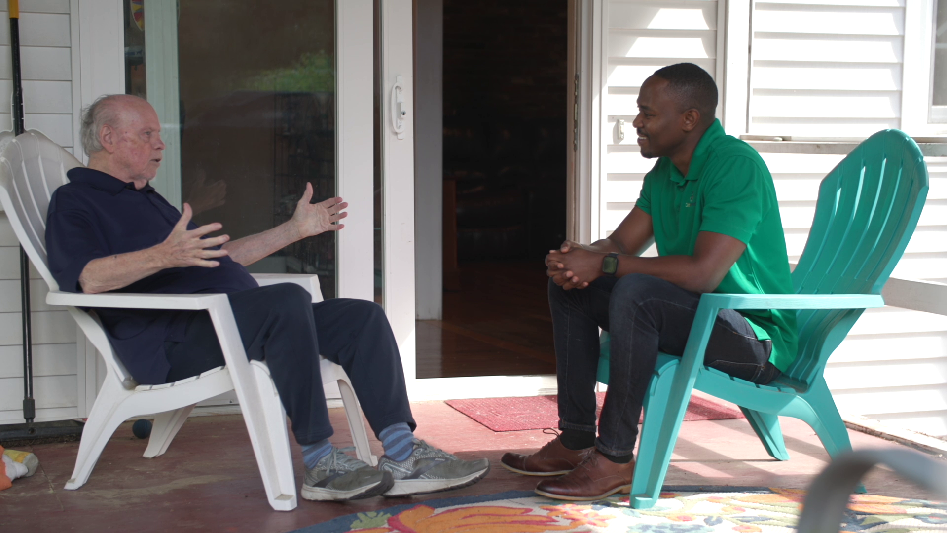 Two people sit on porch chairs, talking across a doorway on a sunny front porch.
