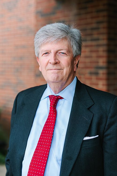 Portrait of a man in a dark suit and red tie standing before a brick wall